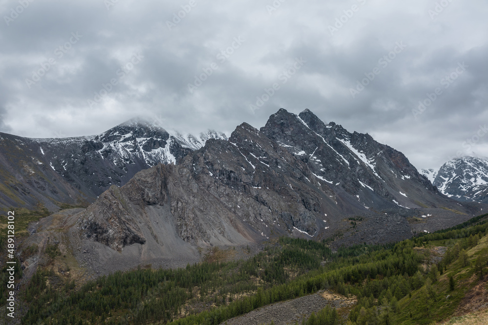 Dark atmospheric landscape with large thorny dragon shaped mountain top under gray cloudy sky. Gloomy aerial view to high snowy mountain range with sharp rocks in shape of dragon back in low clouds.