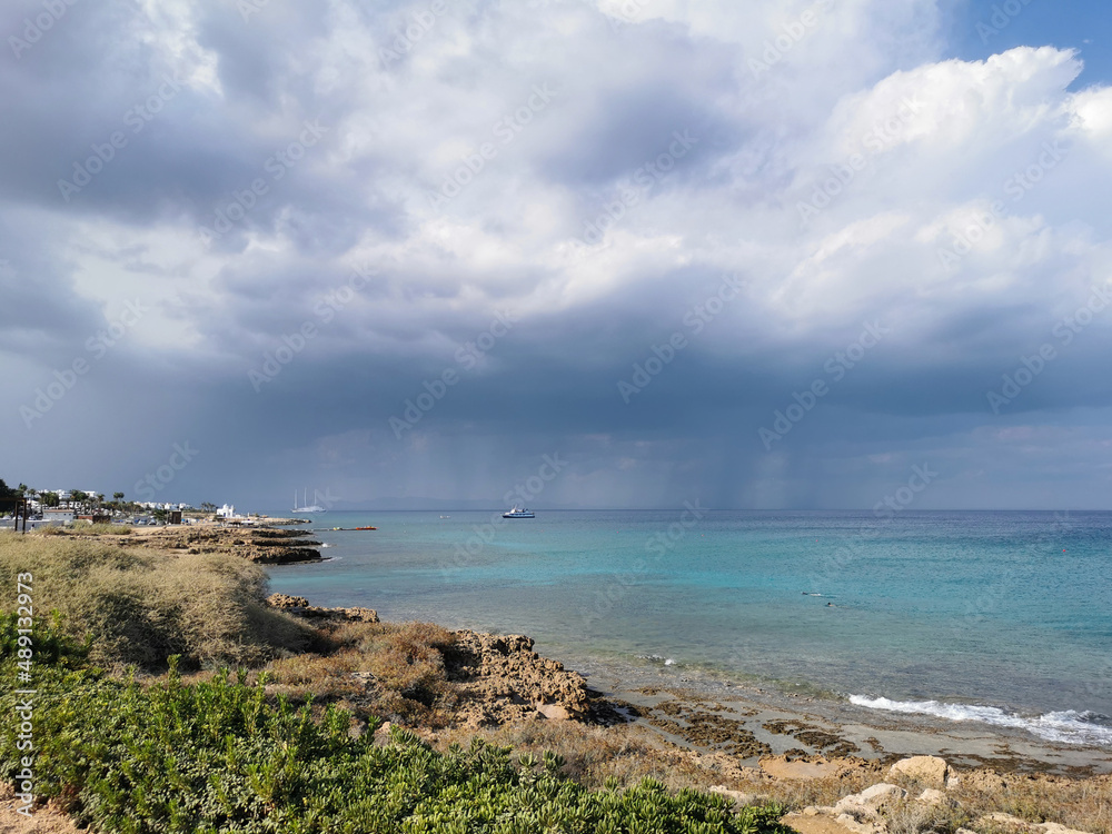 Sunny coast of the Mediterranean Sea with stones, dramatic sky and rain above the sea, boats in the sea and the largest sailing yacht in the world. Sunny coast of the Mediterranean Sea with stones, dramatic sky and rain above the sea, boats in the sea and the largest sailing yacht in the world.
