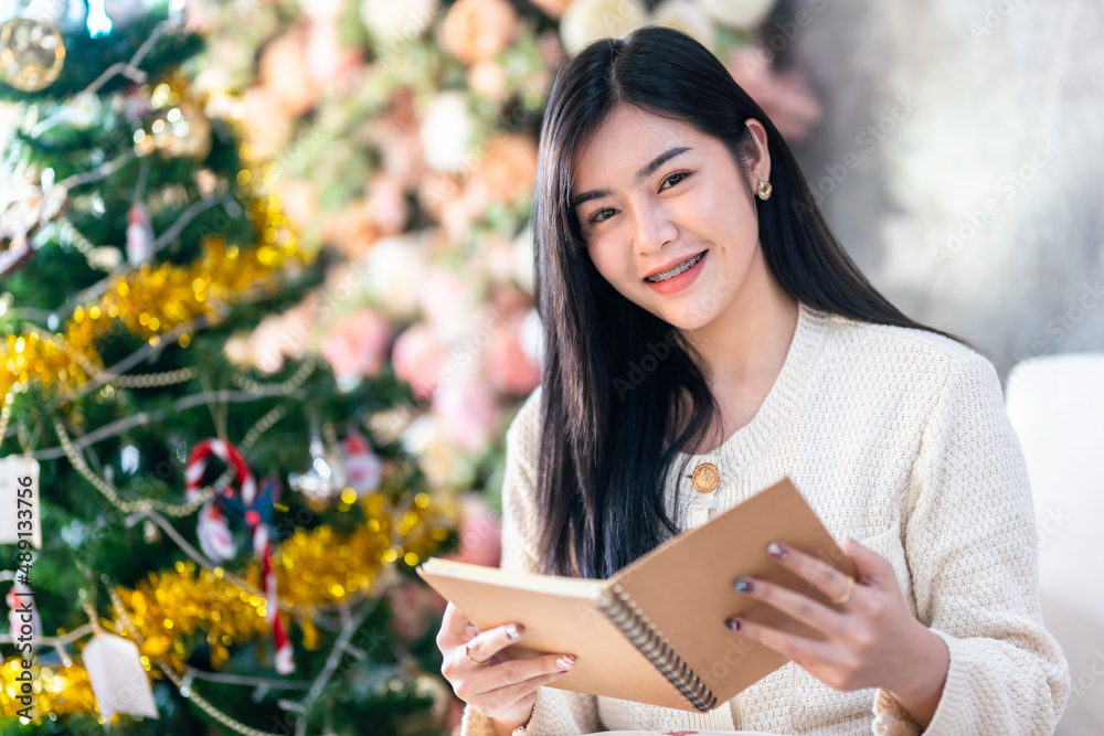 Fototapeta premium Portrait Cute beautiful positive smile young asian woman holding of read a book at home in the living room indoors Decoration During Christmas x-mas and New Year holidays.