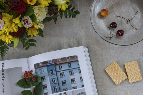 A book with unclear illustrations and photographs of Copenhagen, a bouquet of fresh flowers, appetizing waffles and cherries and apricots on a glass transparent plate on a linen fabric background.