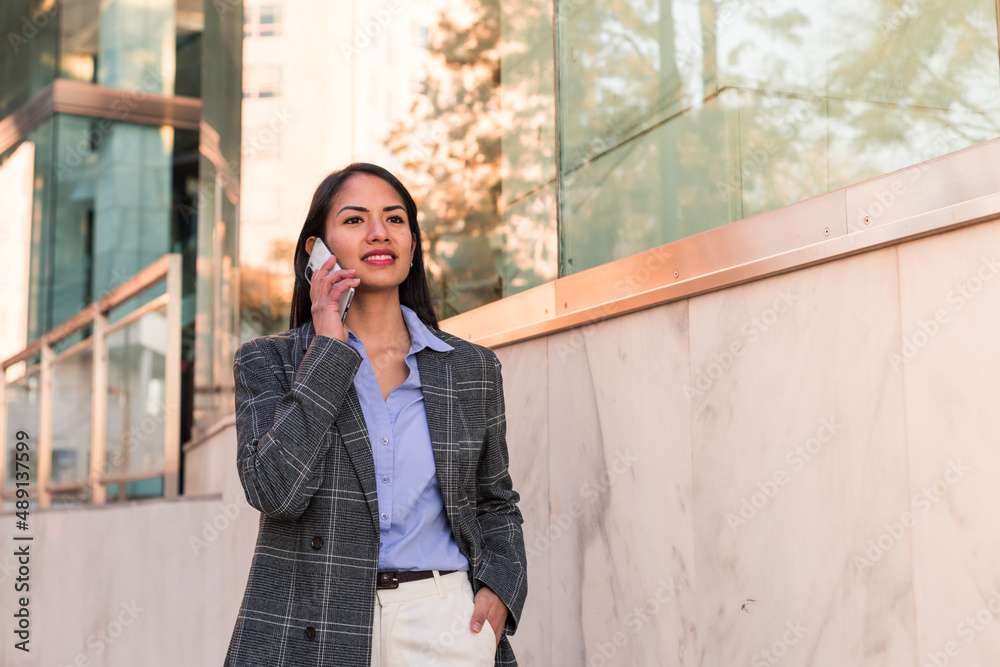Smiling latin american woman talking on phone and looking away wears formal wear near the work place outside. Smiling latin american woman talking on phone and looking away wears formal wear near the work place outside.
