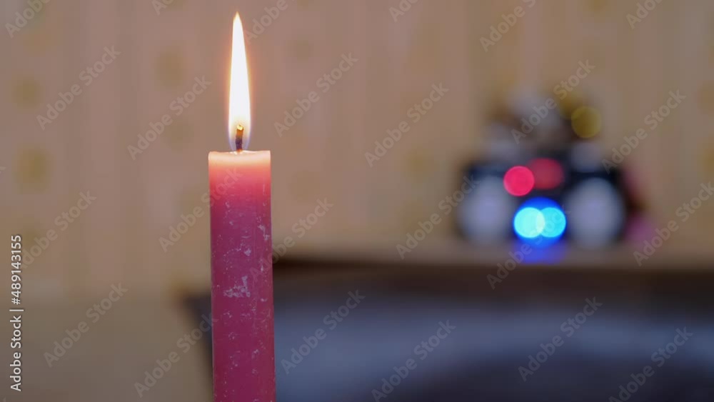 Burning Red Candle with a Bright Moving Flame on a Blurred Background of Lights. One wax, paraffin candle burns in a home interior against background of orange wallpaper. Holiday or memory concept.