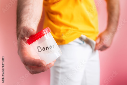 Sperm donation. A man in white jeans zips up his fly and shows a close-up of a plastic jar with sperm samples in his hands. Pink background. The concept of artificial insemination © _KUBE_