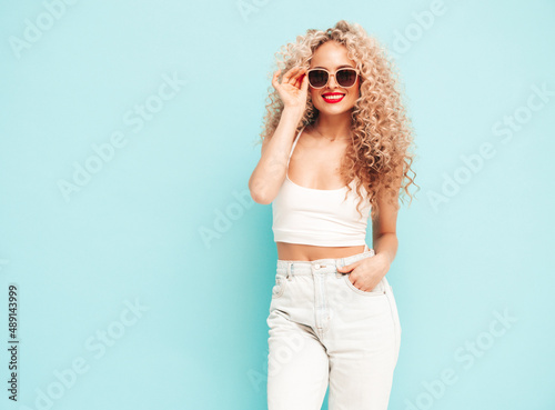 Sexy young beautiful smiling female in trendy summer hipster clothes. carefree woman posing near blue wall in studio. Positive brunette model with afro curls hairstyle. Cheerful and happy