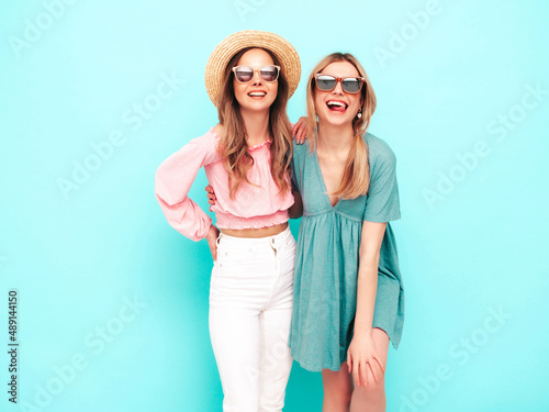 Two young beautiful smiling brunette hipster female in trendy summer dresses. Sexy carefree women posing near blue wall. Positive models having fun. Cheerful and happy. In hats and sunglasses