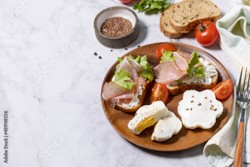 Easter breakfast or brunch. Delicious breakfast or snack - poached egg and cream cheese toast whole grain rye bread, prosciutto, arugula on a marble tabletop. Copy space.