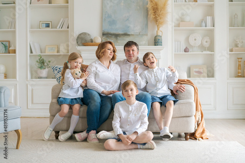 Portrait large family spending time together at home. They are happily sitting in the living room on the couch in a modern light interior. Caucasian parents with children spend time on weekends