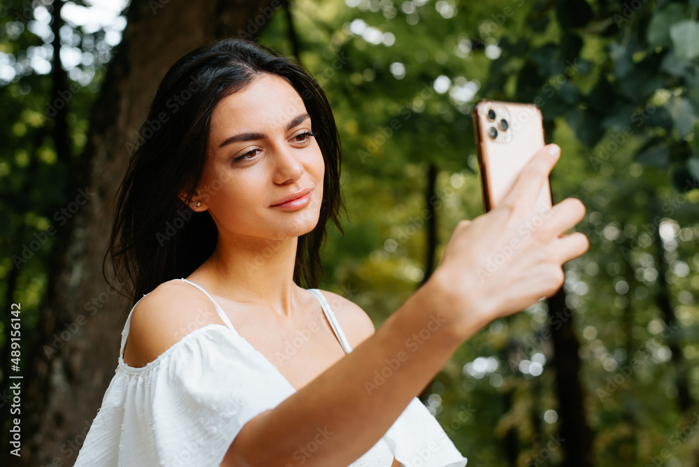 Pretty young mixed race woman taking photo, selfie portrait on mobile phone camera outdoors. Smiling brunette indian lady in white summer dress using smartphone Pretty young mixed race woman taking photo, selfie portrait on mobile phone camera outdoors. Smiling brunette indian lady in white summer dress using smartphone