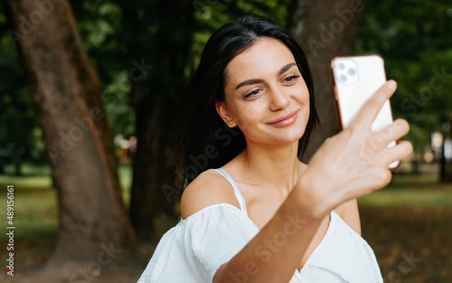 Pretty young mixed race woman with cute smile taking photo, selfie portrait on mobile phone front camera outdoors. Indian brunette lady in white summer dress using smartphone in park during day