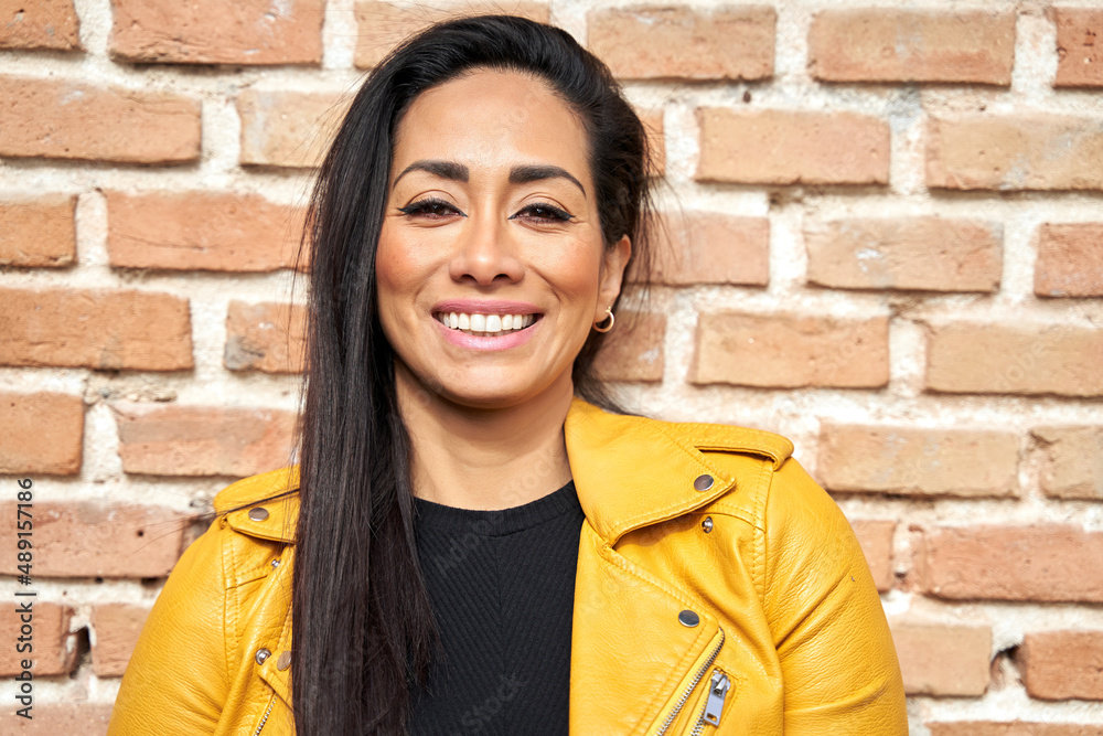 Optimistic Hispanic female with dark hair and in leather jacket standing against brick wall in street and looking at camera Optimistic Hispanic female with dark hair and in leather jacket standing against brick wall in street and looking at camera