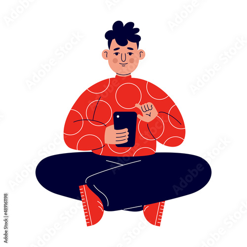 A young man in a red sweater sits on the floor and works with the phone on social networks. (ID: 489161198)