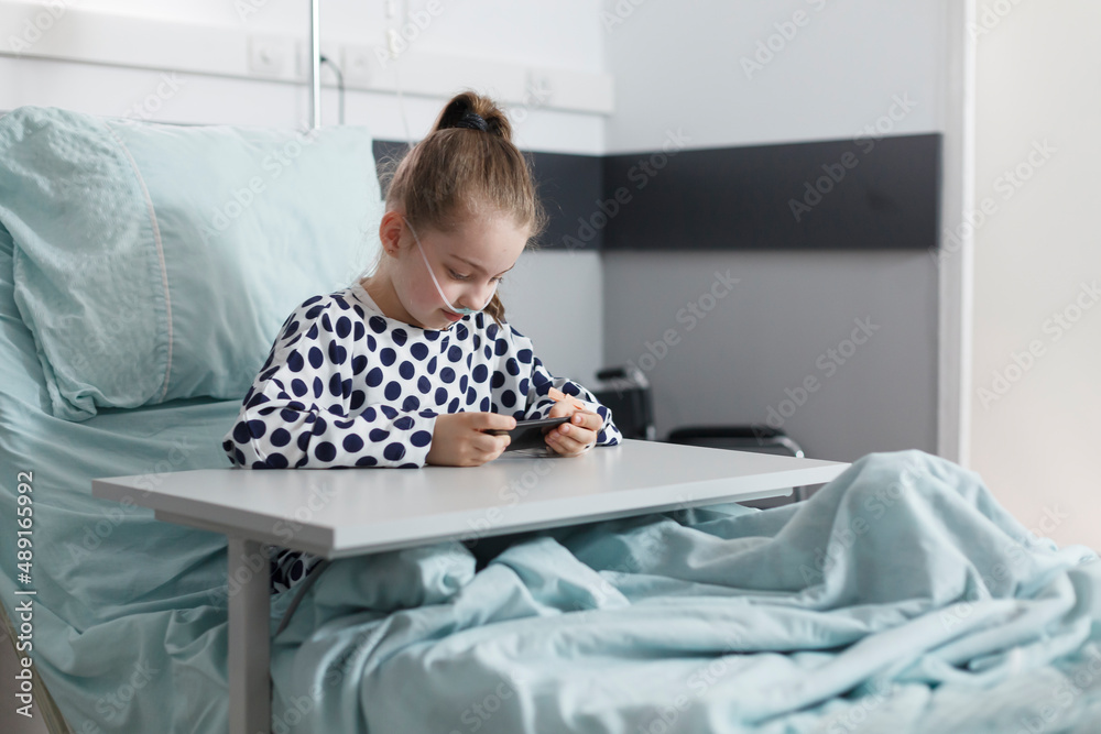 Sick little girl playing on smartphone while resting on patient bed in pediatric clinic room. Ill young patient enjoying mobile phone entertainment while sitting in hospital recovery ward. Sick little girl playing on smartphone while resting on patient bed in pediatric clinic room. Ill young patient enjoying mobile phone entertainment while sitting in hospital recovery ward.