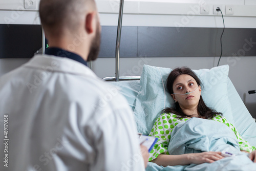 Over shoulder view of woman recieving oxygen recovering in hospital bed looking at doctor holding clipboard with lab results. Patient with nasal cannula with low spo2 saturation in hospital ward.