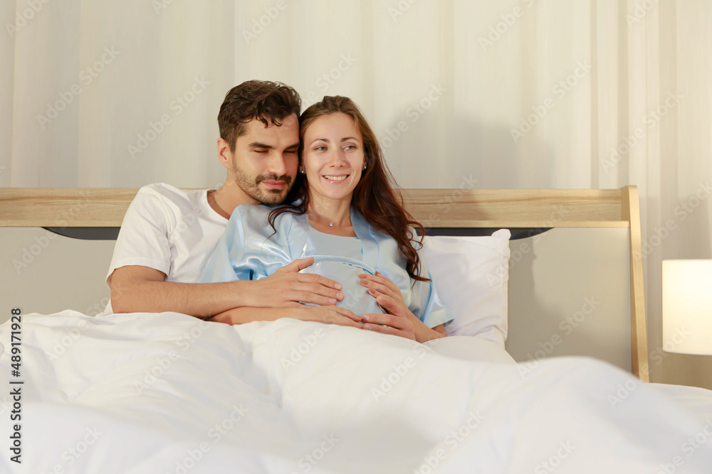 Fototapeta premium Cheerful husband enjoy embracing pregnant wife and happy to tenderly fondle beloved belly of unborn baby and expecting warm family during prenatal care of maternity at romantic bedroom