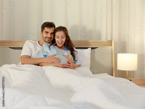 Cheerful husband enjoy embracing pregnant wife and happy to tenderly fondle beloved belly of unborn baby and expecting warm family during prenatal care of maternity at romantic bedroom