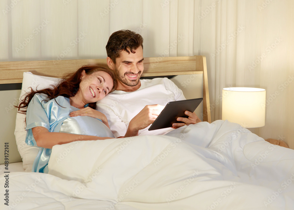 Fototapeta premium Happy pregnant wife holding belly and relax on bed beside beloved husband and enjoy watching entertainment media on tablet together with smile and laugh. Parents expecting joyful life for child birth.
