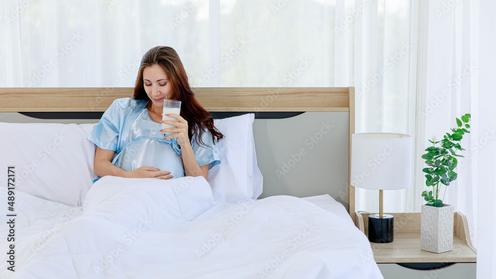 Cheeful pregnant woman smile as enjoy drinking glass of milk beverage in morning while resting on bed with lovely bear doll