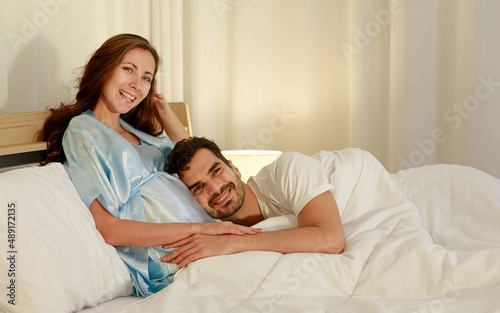 Happy Caucasian husband tenderly lies down on belly and embrace lovely pregnant wife who smile on bed