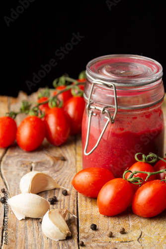Top view of homemade ketchup jar on rustic wooden table with pepper, garlic and cherry tomatoes, selective focus, black background, vertical