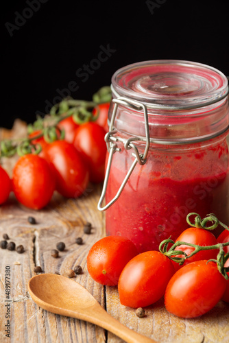 Homemade ketchup jar closeup on rustic wooden table with spoon, pepper and cherry tomatoes , selective focus, black background, vertical