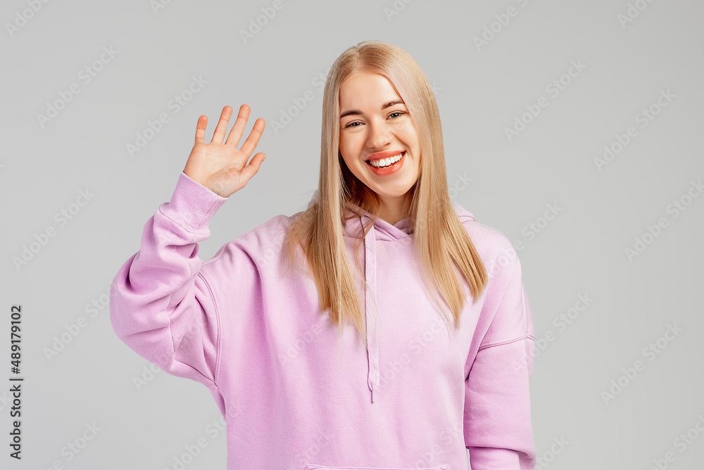 Portrait of smiling happy girl waving friendly, saying hello, greeting, hi gesture, standing in violet hoodie over gray background
