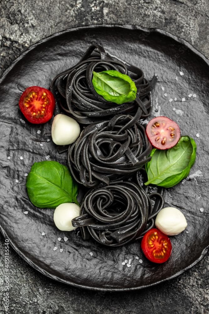 black pasta caprese salad tomatoes, mozzarella and green basil leaves. Pasta with cuttlefish ink. Vegan or gluten free diet. vertical image. top view. place for text