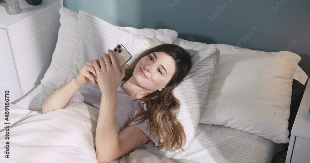 Beautiful caucasian woman lying in bed, click and swipe on phone. Happy attractive female spend leisure time at home, feel relax and enjoy communicate on smartphone.