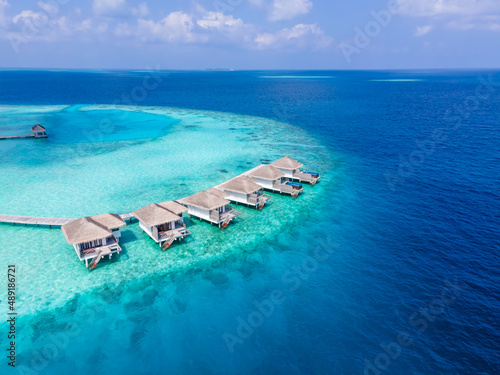 Overwater villas on tropical atoll island for holidays vacation travel and honeymoon. Luxury resort hotel in Maldives or Caribbean with turquoise sea water. Drone aerial view.