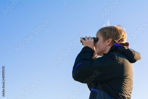 Blonde young man looking through binoculars while outdoors in nature. Guy looking through binoculars on the blue sky. Travel and wildlife concept