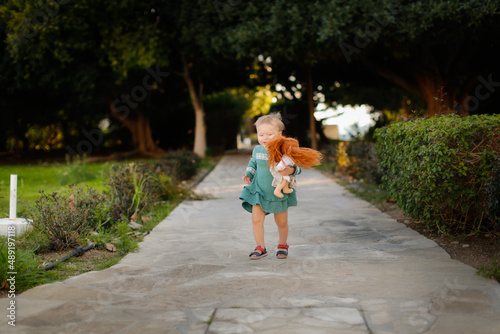 Cute european baby toddler girl in a dress with a doll runs walks in the park. One child on the path on grass in the park © natalialeb