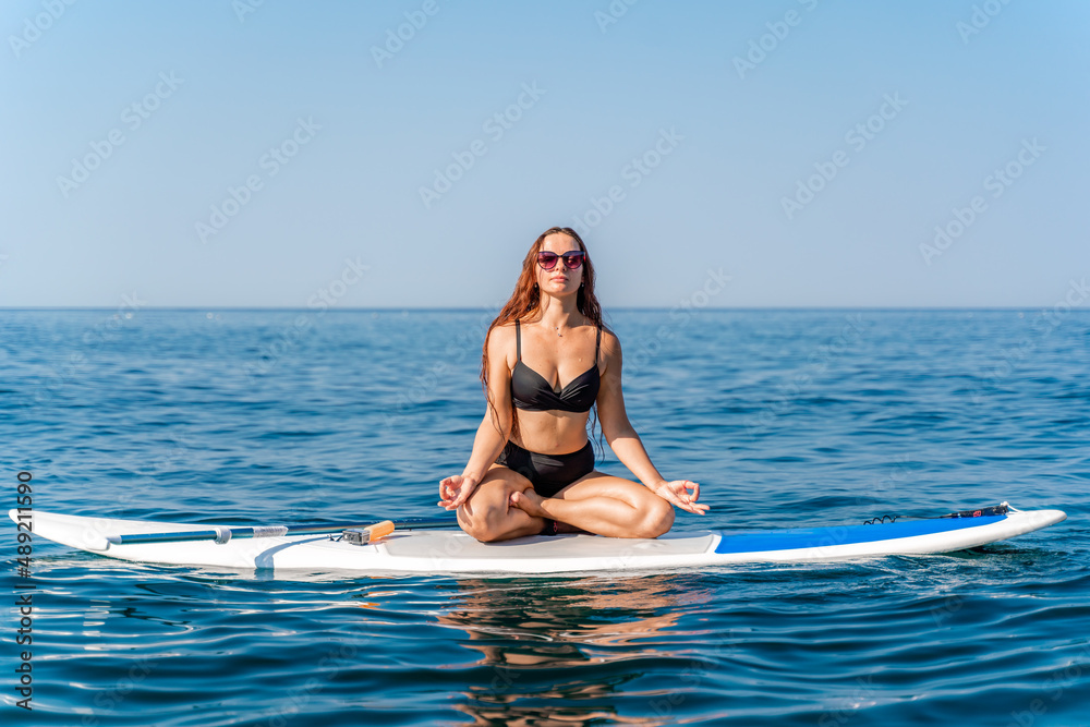 Sporty girl on a glanders surfboard in the sea on a sunny summer day. In a striped swimsuit, he sits in the lotus position with his eyes closed.