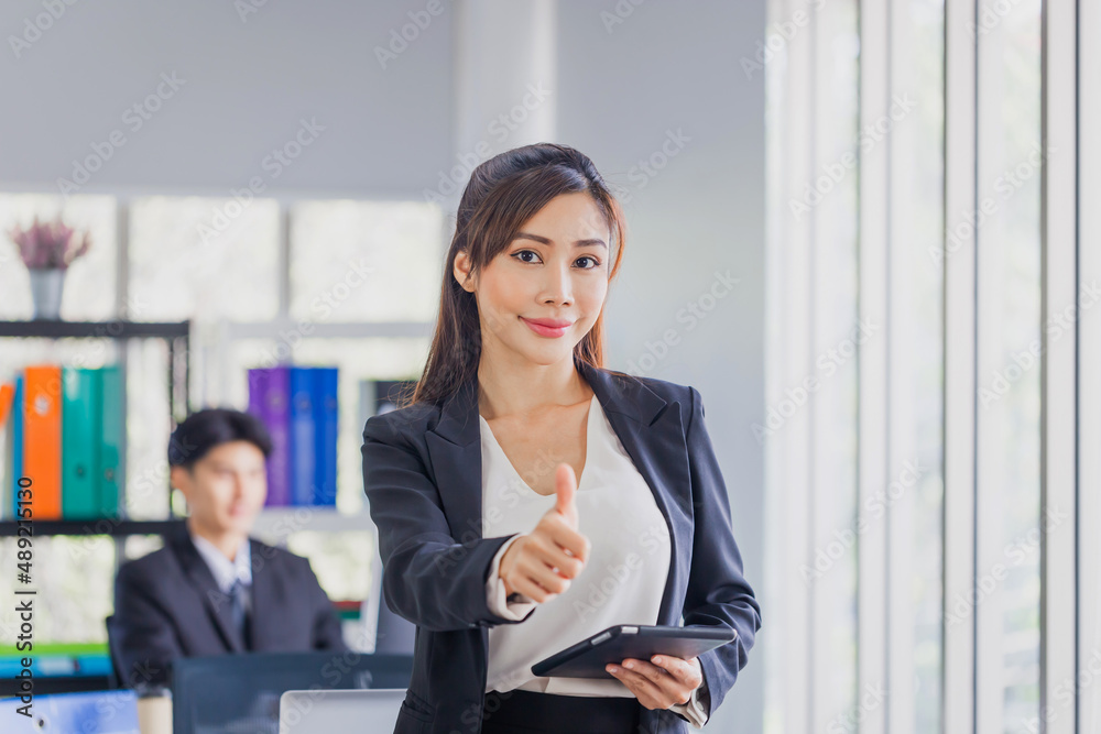Attractive cheerful young business woman showing thumbs up and looking at camera and blurred colleague sitting in the background Attractive cheerful young business woman showing thumbs up and looking at camera and blurred colleague sitting in the background