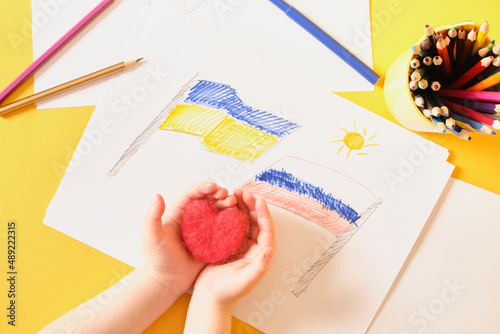 children's drawing, flags of russia and ukraine are drawn on a sheet, children's hands hold a heart made of felt