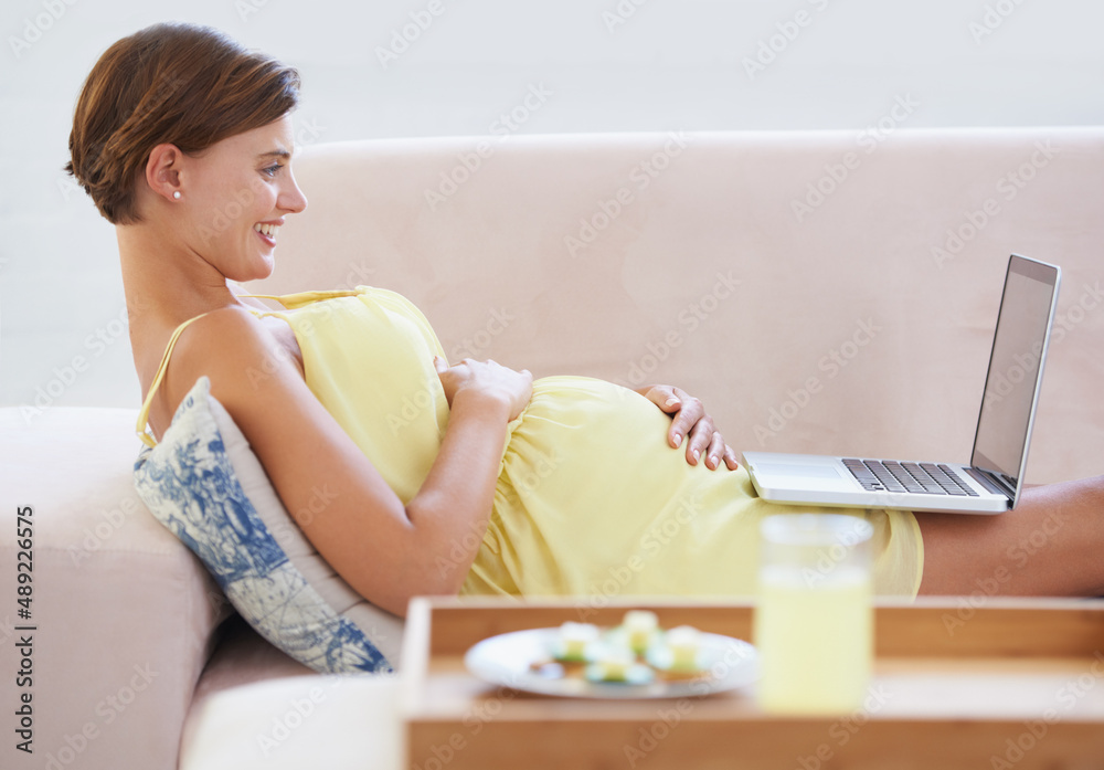 In good spirits. A smiling pregnant woman lying on the couch using a laptop with a healthy meal beside her. In good spirits. A smiling pregnant woman lying on the couch using a laptop with a healthy meal beside her.