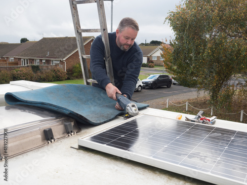 A man can be seen working from a ladder using an angle grinder to remove a solar panel from the roof top of a motorhome recreational vehicle.Viewed at roof top height