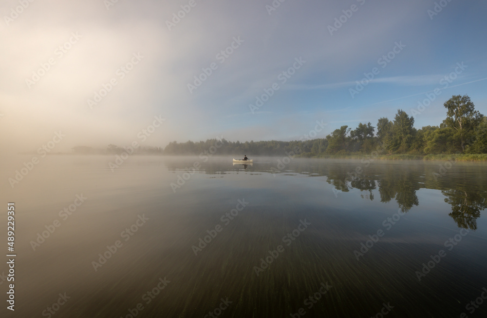 Fototapeta premium Silhouette of a man in a boat. Mystical landscape. Fog in the early morning on the river. The trees near the water are illuminated by the rays of the rising sun.