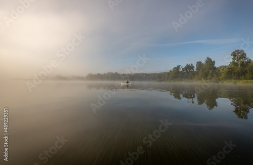 Fototapeta Naklejka Na Ścianę i Meble -  Silhouette of a man in a boat. Mystical landscape. Fog in the early morning on the river. The trees near the water are illuminated by the rays of the rising sun.