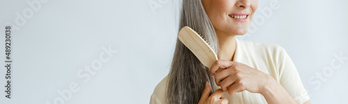 Thoughtful middle aged Asian woman brushes loose grey hair posing on light background in studio, space for text. Mature beauty lifestyle