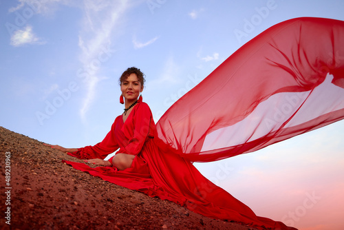 Beautiful young woman or girl with curly hair and in red dress with a light flying fabric on the sand on sunny day with blue sky in the background. Model or dancer posing in photo shoot on sand dunes
