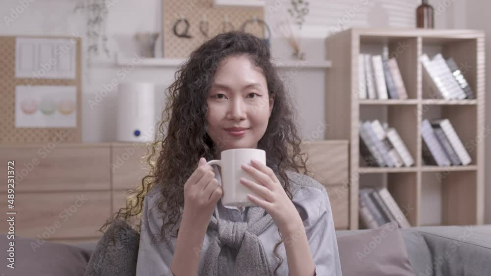 Asian Woman pondering at home in her leisure time while reclining on the sofa and drinking hot tea. On a lovely winter morning, a joyful girl relaxes at home.