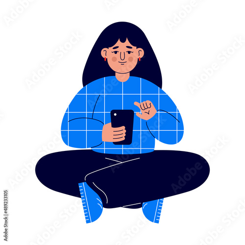 A young woman in a blue sweater sits on the floor and works with the phone on social networks. (ID: 489233105)