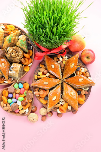 Fototapeta Naklejka Na Ścianę i Meble -  Nowruz festive table. green wheat grass with red ribbon, arabic dessert baklava, sweets, nuts, dry fruits on pink background. Spring equinox in March, Nowruz Holiday. top view