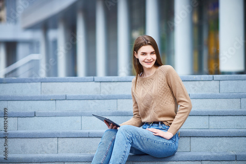 young girl sits on the steps on the street with a tablet in her hands and looks at the camera, smiling. Copy space © Татьяна Кутина