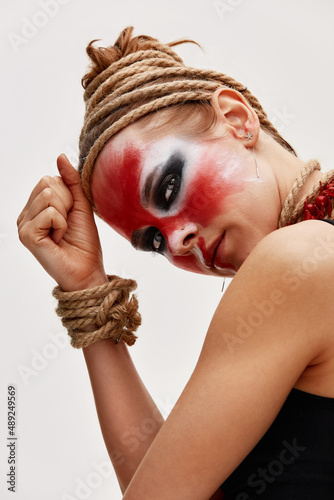 close up portrait of young beautiful girl with colorful face painting. Halloween professional makeup. hair in paint. neck and hands are wrapped with ropes. Victim Concept