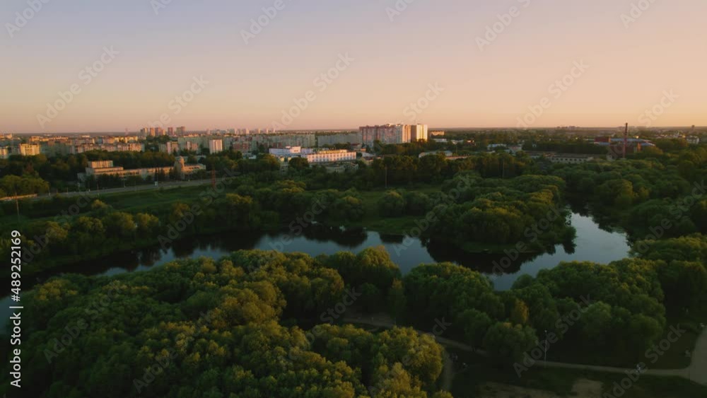 Smooth aerial flight over the evening city at sunset. A cityscape overview of the park, the river and the houses behind. Romantic bird's eye view of the town during sundown time and golden hour.