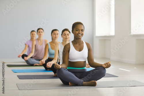 Portrait of happy young african american woman relaxing in group yoga class in studio. Woman sits on mat in lotus position among other meditating caucasian women and looks at camera. Yoga concept.
