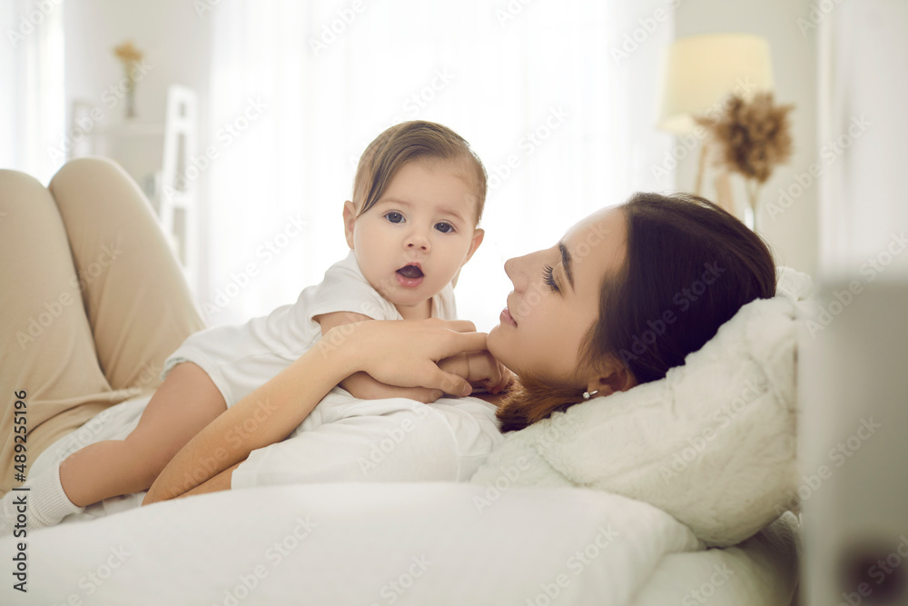 Happy mother together with her child cuddling on comfy bed at home. Young mommy and adorable baby in comfortable white cotton bodysuit lying on bed in the morning or afternoon. Family and care concept Happy mother together with her child cuddling on comfy bed at home. Young mommy and adorable baby in comfortable white cotton bodysuit lying on bed in the morning or afternoon. Family and care concept