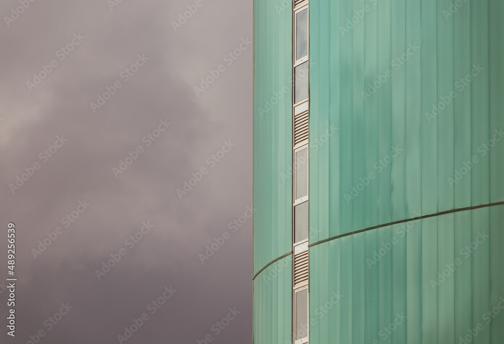 View of the green facade of a tall modern building with a vertical row of narrow windows and the cloudy sky on the left View of the green facade of a tall modern building with a vertical row of narrow windows and the cloudy sky on the left