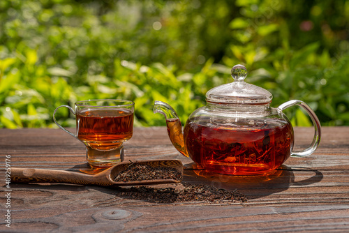 Steamed glass teapot, elegant cup. Bamboo spoon with tea, wooden table. Outdoor, picnic, brunch. Floral background in blur.