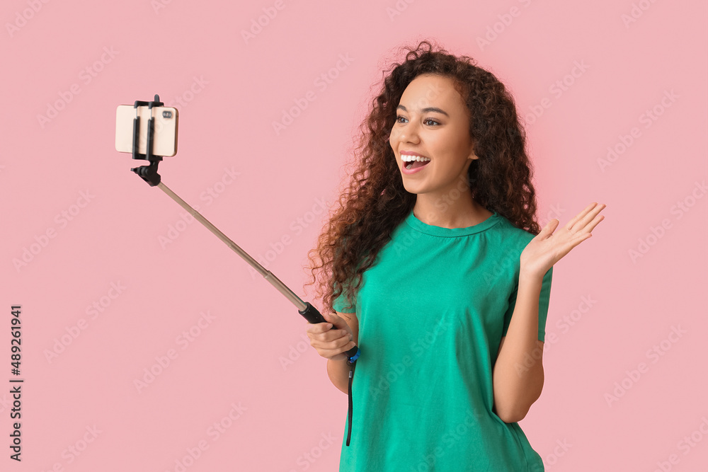 Young African-American woman with mobile phone taking selfie on pink background Young African-American woman with mobile phone taking selfie on pink background